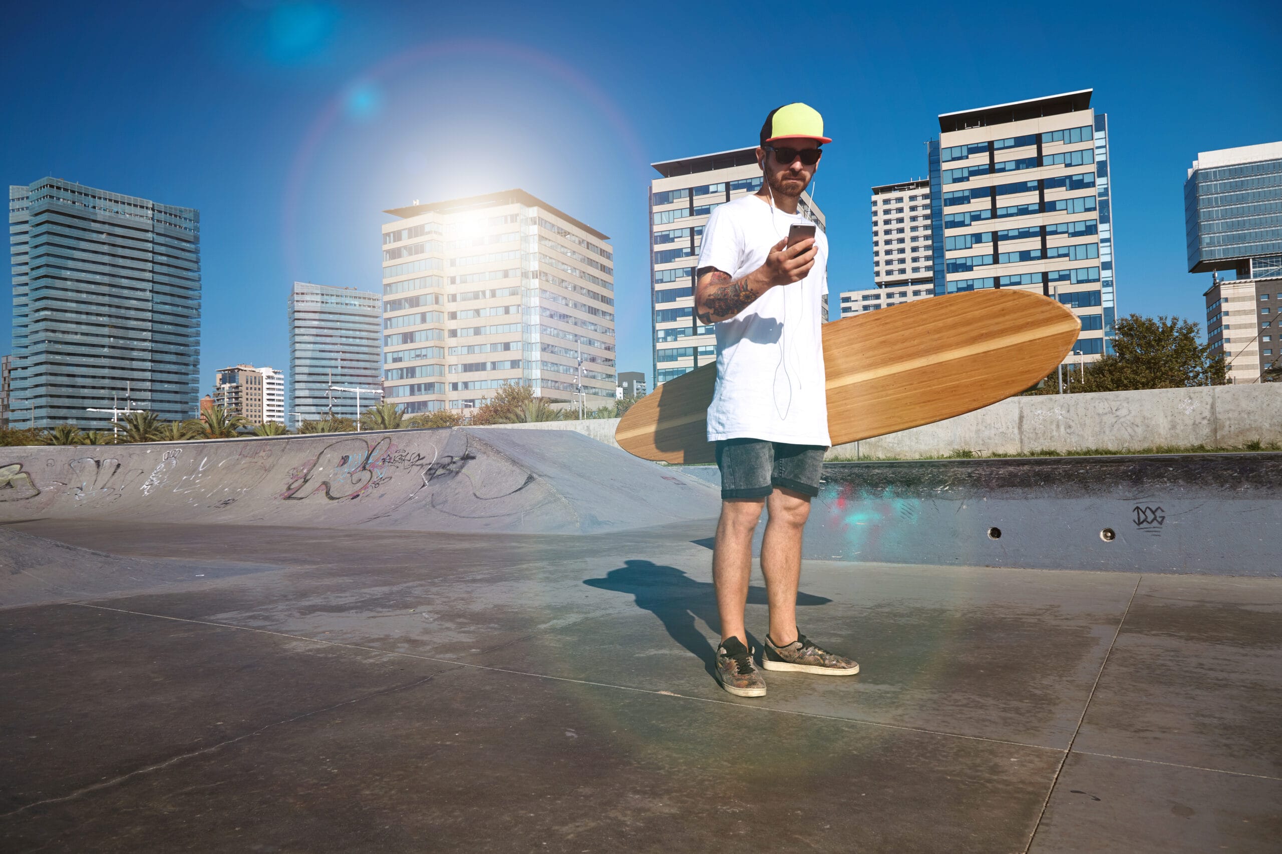 Young attractive bearded man with tattooes wearing unlabeled white t-shirt looks at his smartphone ,holds big bamboo longboard in center of concrete pool in urban skatepark